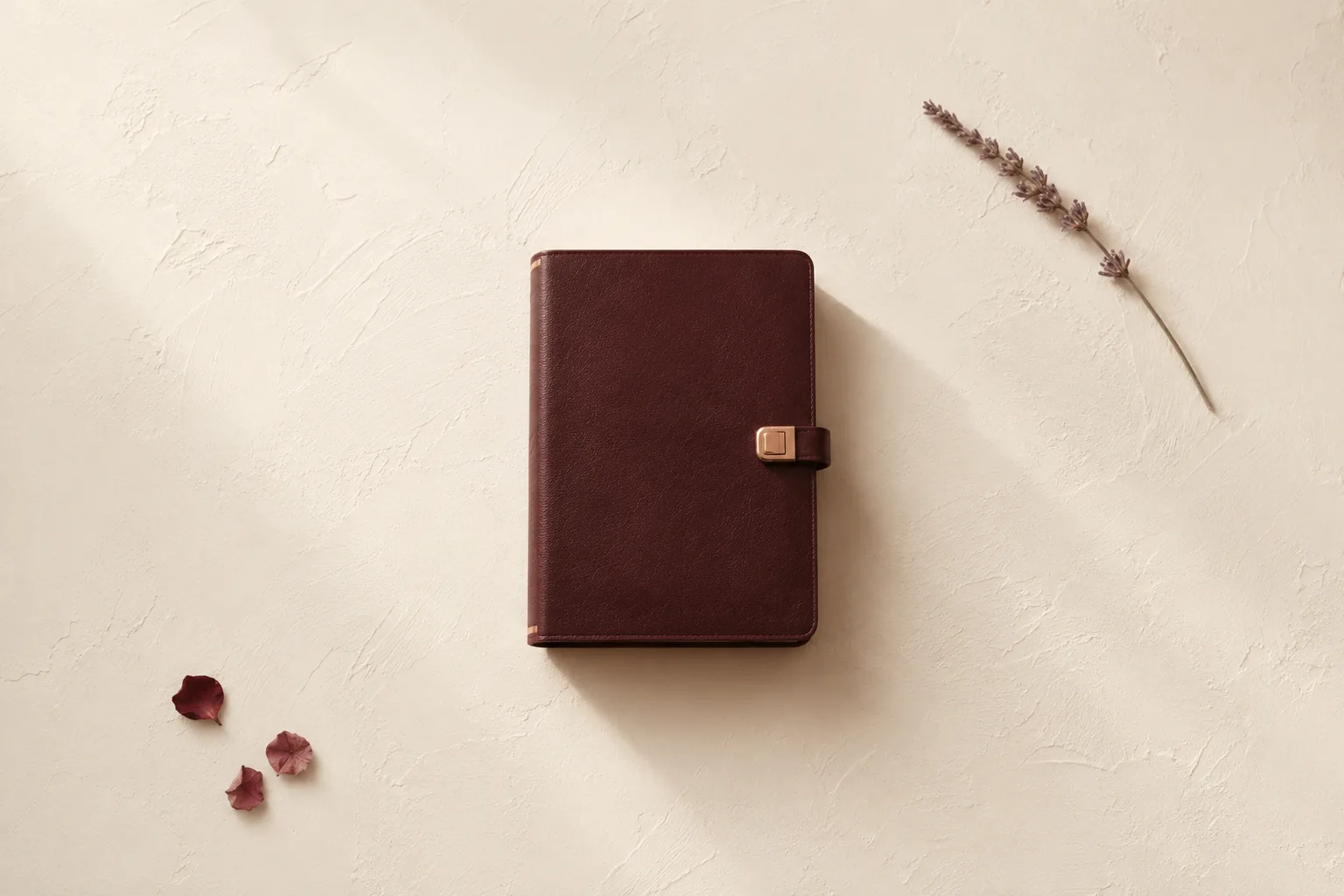 A closed burgundy leather journal with a small rose-gold clasp lock, alone on a cream plaster surface